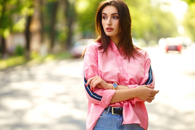 Porträt der jungen und schönen Frau in der Freizeitkleidung auf der Straße. gekleidet in ein rosa Hemd und Jeans. Frühjahr / Sommer-Konzept. Zeit zum Entspannen. Mädchen mit blauen Augen. Genießen Sie den Sonnenuntergang auf der Straße