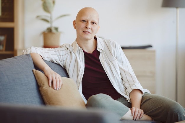 Porträt der kahlen erwachsenen Frau, die Kamera beim Sitzen auf der Couch im modernen Hauptinnenraum, Alopezie und Krebsbewusstsein, Kopierraum betrachtet