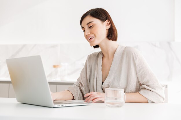 Porträt der kaukasischen erwachsenen Frau, die Robe lächelnd und arbeitend trägt oder auf weißem Laptop in Wohnung plaudert