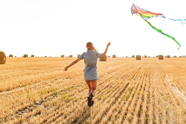 Porträt der kaukasischen Frau 20s lächelnd und spielend mit fliegendem Drachen während des Spaziergangs durch goldenes Feld, während des sonnigen Tages