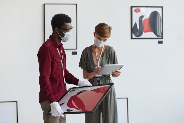 Porträt der kreativen tätowierten Frau unter Verwendung des digitalen Tablets während der Planung der Kunstgalerie-Ausstellung mit afroamerikanischem Assistenten,