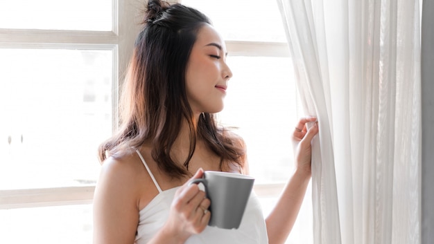 Porträt der lächelnden glücklichen fröhlichen schönen hübschen asiatischen Frau, die das Trinken und das Betrachten der Tasse heißen Kaffees oder des Tees entspannt. Mädchen, das gefällt, genießt das Frühstücken im Ferienmorgenurlaub auf dem Bett zu Hause