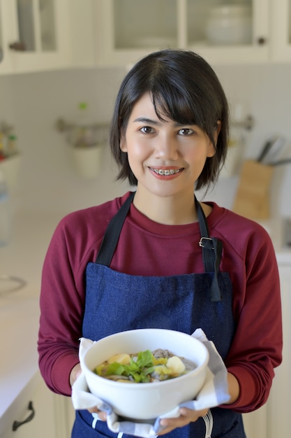 Porträt der lächelnden jungen Frau in der modernen Küche und im Halten einer Schüssel Suppe mit Tofu