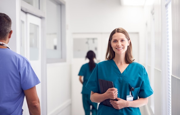 Porträt der lächelnden Ärztin, die Scrubs im beschäftigten Krankenhaus-Korridor trägt, der Digital-Tablet hält