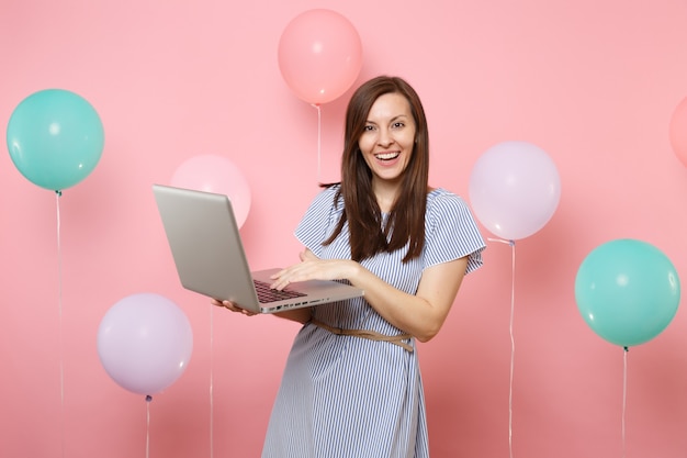 Porträt der lächelnden schönen jungen Frau im blauen Kleid, die unter Verwendung des Laptop-PC-Computers auf pastellrosa Hintergrund mit bunten Luftballons hält. Geburtstagsfeier, Konzept der aufrichtigen Emotionen der Menschen.