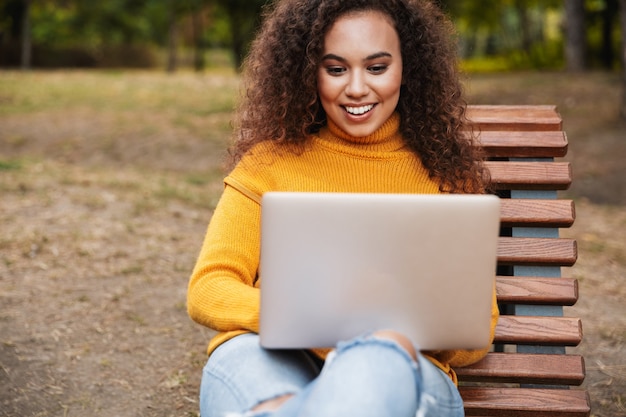 Porträt der lächelnden schönen jungen gelockten Frau im Park draußen unter Verwendung der Laptop-Computer.