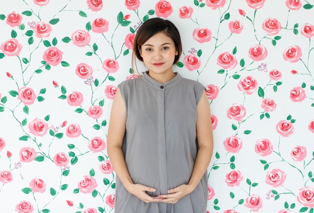 Porträt der lächelnden Stellung der jungen asiatischen schwangeren Frau, die ihren Bauch berührt, der eine Kamera über weißem Rosenmusterhintergrund betrachtet. Gesundes und glückliches Damenkonzept.