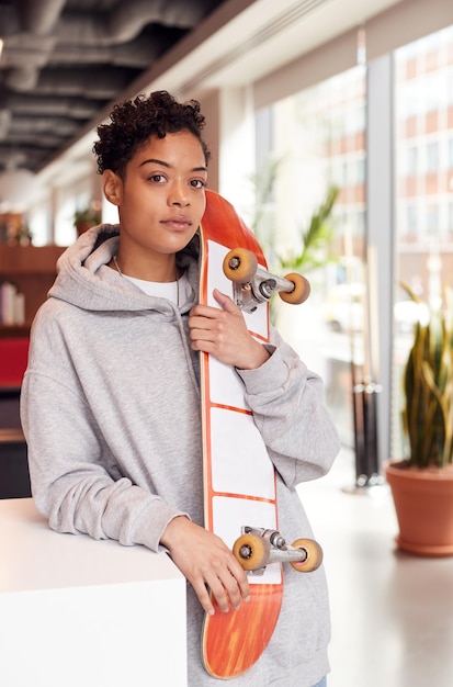 Porträt der lässig gekleideten jungen Geschäftsfrau mit dem Skateboard, das im Großraumarbeitsplatz steht