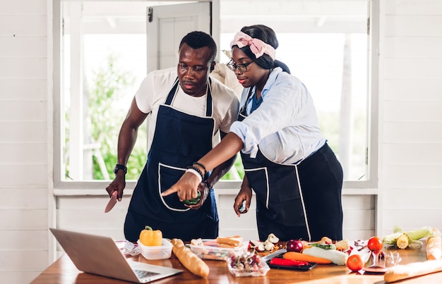 Porträt der Liebe afroamerikanisches Paar, das Spaß hat, zusammen zu kochen und nach Rezept im Internet mit Laptop-Computer zu suchen, um das leckere Essen in der Küche zu Hause vorzubereiten