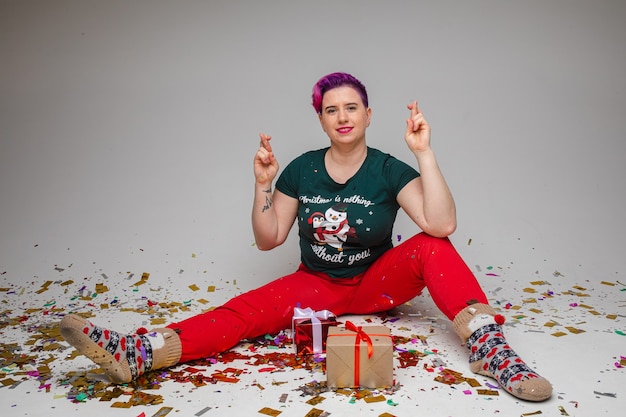 Porträt der lustigen gemütlichen frau mit lila haaren im weihnachtspyjama und in den socken, die zwei verpackte geschenke in den händen zeigen, während sie auf dem boden sitzen, der mit konfetti bedeckt ist