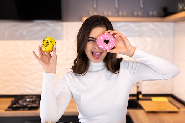 Porträt der lustigen jungen Frau, die ihre Augen mit Donuts in der modernen Küche bedeckt.