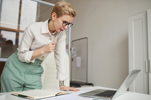 Porträt der modernen Geschäftsfrau, die über Schreibtisch beim Betrachten des Laptop-Bildschirms im Büro, Kopierraum verlässt