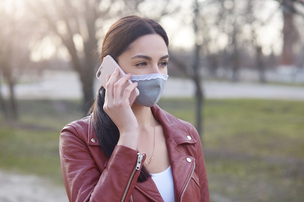 Porträt der nachdenklichen ernsten Brünette, die Smartphone nahe Ohr hält, das medizinische Maske trägt