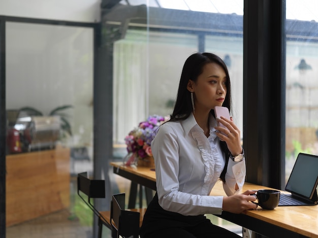 Porträt der nachdenklichen Frau, die Smartphone und Kaffeetasse beim Sitzen am Arbeitsplatz hält