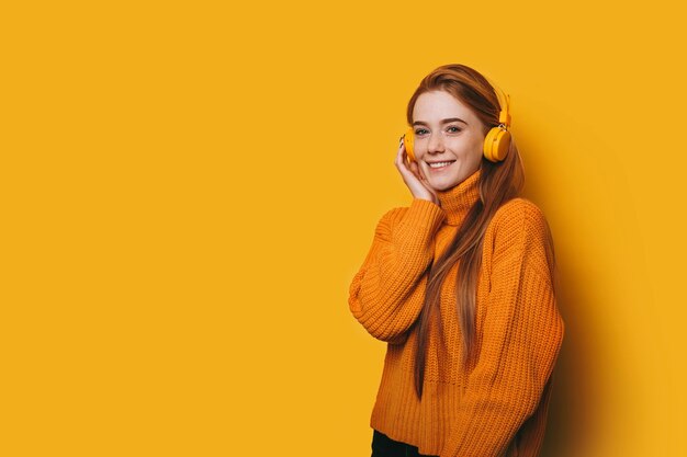 Porträt der niedlichen jungen Frau mit den roten Haaren und den Sommersprossen, die Kamera lächelnd betrachten, während Musik auf gelben Kopfhörern gegen gelbe Wand hören.