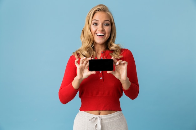 Porträt der optimistischen blonden Frau 20s, die rotes Hemd trägt, das lächelt und leeren Bildschirm des Handys zeigt, lokalisiert über blaue Wand