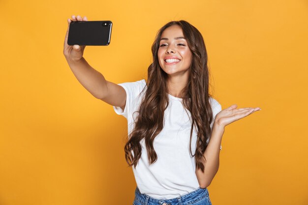 Porträt der positiven Frau 20s mit langen Haaren, die lachen, während Selfie-Foto auf Smartphone nehmen, lokalisiert über gelbe Wand