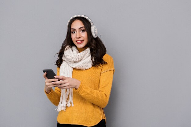 Porträt der positiven Frau, die Ohrenschützer und Schal hält Handy trägt, lokalisiert über grauem Hintergrund