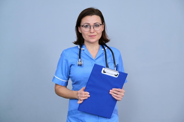 Porträt der reifen Krankenschwesterfrau, die Zwischenablage hält. Weibchen in blauer Uniform mit Stethoskop, das auf grauem Hintergrund in die Kamera schaut