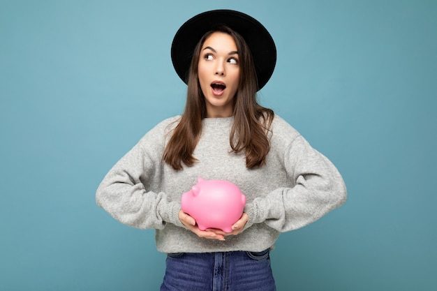 Porträt der schockierten verblüfften jungen schönen schönen brünetten Frau, die stilvollen grauen Pullover und schwarzen Hut lokalisiert über blauem Hintergrund mit freiem Raum trägt und rosa Schweinchenbox hält. Sparbüchsenkonzept.