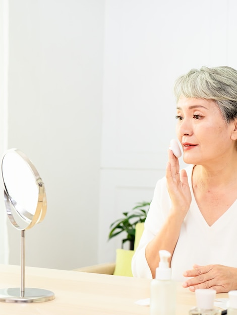 Porträt der schönen älteren asiatischen Frau, die Puderquaste am Backenmake-up der Kosmetik anwendet.