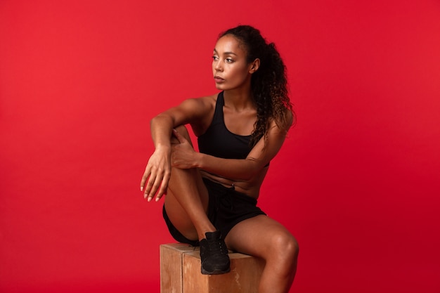 Porträt der schönen Afroamerikanerfrau in der schwarzen Sportbekleidung, die auf Kasten sitzt, lokalisiert über roter Wand
