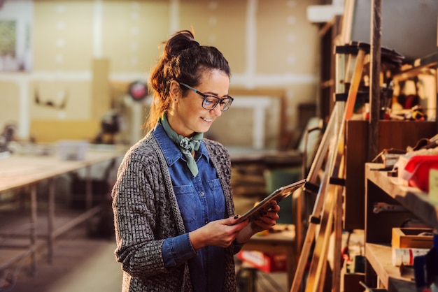 Porträt der schönen Arbeiterin des Mittelalters Industrie, die Tablette in ihren Händen hält und vor Regal mit Werkzeugen steht.