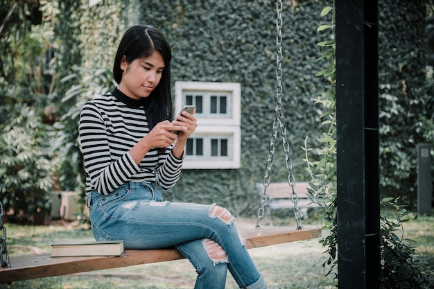 Porträt der schönen asiatischen Frau, die ihren Handy verwendet und auf hölzernem Schwingen unter Baum sitzt.