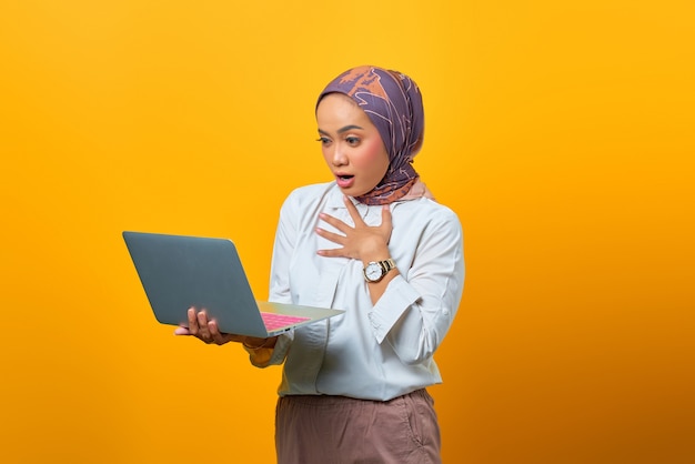 Porträt der schönen asiatischen Frau, die Laptop mit überraschtem Ausdruck über gelbem Hintergrund hält