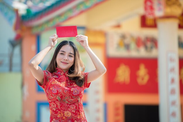 Porträt der schönen asiatischen Frau im Cheongsam-KleidThailänderFröhliches chinesisches NeujahrskonzeptHolding roten Umschlag rot