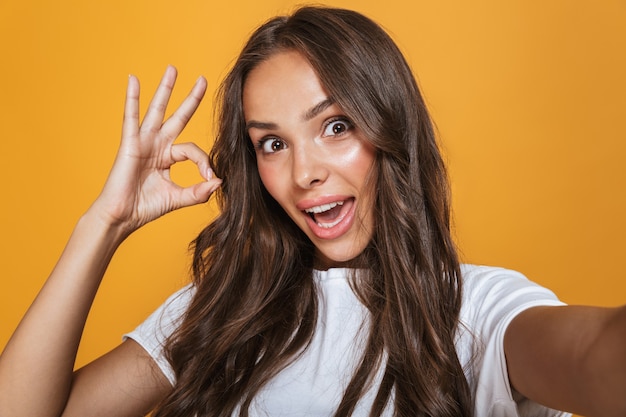 Porträt der schönen Frau 20s mit langen braunen Haaren, die lachen, während sie Selfie-Foto machen, lokalisiert über gelber Wand