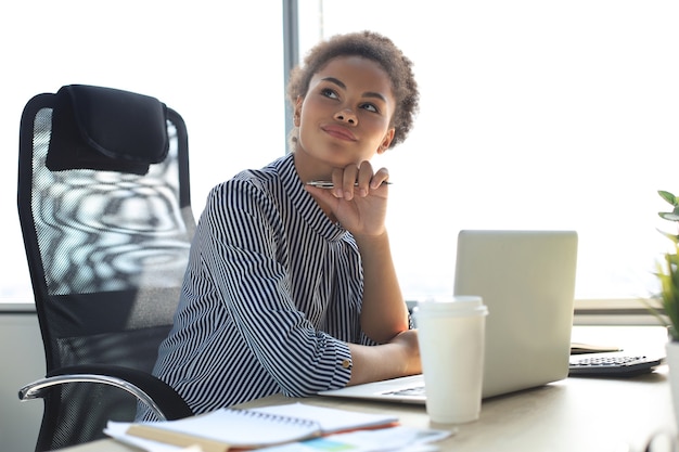 Porträt der schönen jungen Afroamerikanerfrau, die mit Laptop beim Sitzen am Tisch arbeitet.