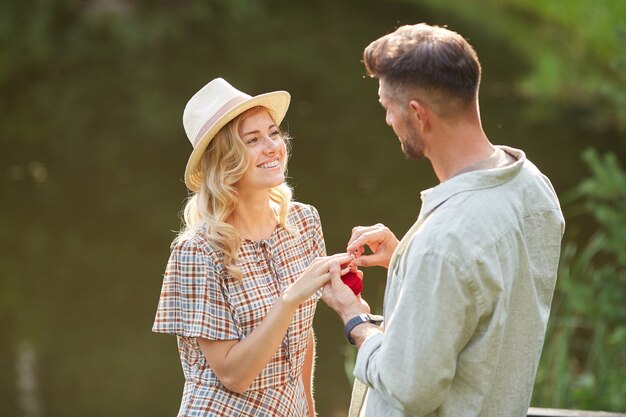 Porträt der schönen jungen Frau, die Heiratsantrag annimmt, während am See im Sonnenlicht steht