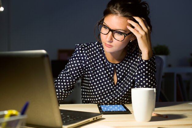 Porträt der schönen jungen Frau, die nachts mit Laptop in ihrem Büro arbeitet.