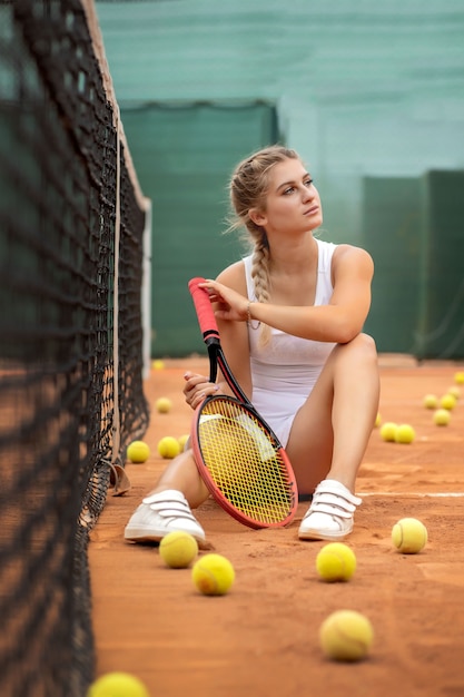 Porträt der schönen jungen Frau, die nahe Netz im Tennisplatz mit Ball im Freien sitzt. Überzeugte Sportlerin, die auf Tennisplatz stillsteht.