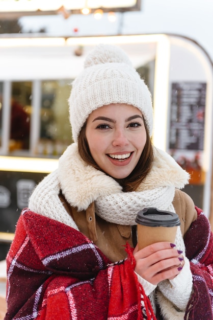 Porträt der schönen jungen hübschen Frau im Hut und im Schal, die draußen in der schneebedeckten Parkstraße gehen, die Kaffee hält Tasse trinkt.