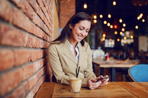 Porträt der schönen kaukasischen Brünette gekleidet klug lässig im Café sitzen und Smartphone für das Senden von Nachricht verwenden. Auf dem Holztisch steht frischer leckerer Kaffee.