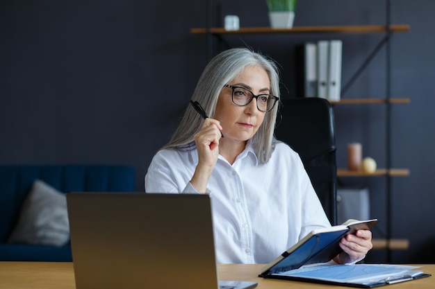 Porträt der schönen lächelnden älteren Frau, die online studiert. Online-Bildung, Fernarbeit, Heimunterricht. Grauhaarige ältere Frau, die im Notizbuch schreibt. Ältere Menschen studieren Online-Kurse.