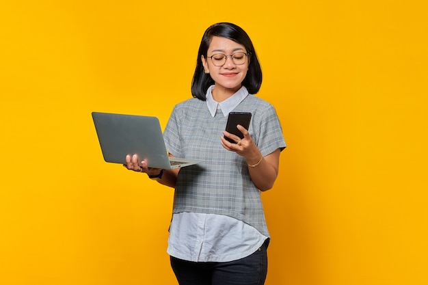 Porträt der schönen lächelnden asiatischen Frau, die Laptop und Handy auf blauem Hintergrund hält