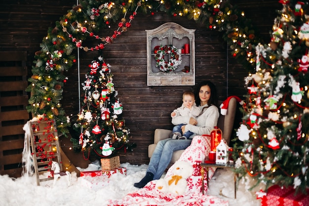 Porträt der schönen lächelnden Frau mit Frisur und Make-up, die ihr reizendes Baby gegen geschmückten Weihnachtsbaum halten