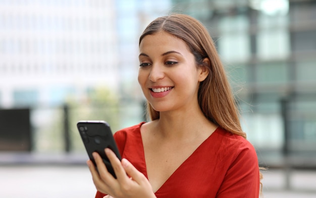 Porträt der schönen lässigen Geschäftsfrau, die Nachricht mit Handy-Chat-App mit modernen Wolkenkratzern auf dem Hintergrund sendet