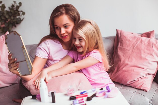 Porträt der schönen Mutter und ihrer entzückenden kleinen Tochter, beide in rosafarbenen T-Shirts und stylischen Stirnbändern, die auf einer Couch sitzen und Lippen färben, die in kleine Tischspiegel schauen