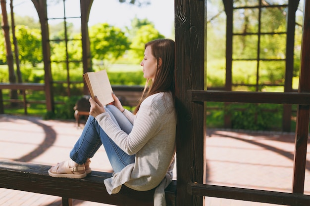 Porträt der schönen ruhigen friedlichen jungen Frau, die leichte Freizeitkleidung, Jeans trägt, sich entspannt, Buch liest. Lächelnde Frau, die im Stadtpark in der Straße draußen auf Frühlingsnatur stillsteht. Lifestyle-Konzept.