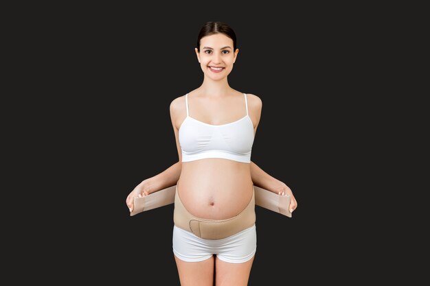 Porträt der schwangeren Frau im Unterwäscheverband Mutterschaftsgürtel gegen Schmerzen im Rücken an der schwarzen Oberfläche mit Kopienraum
