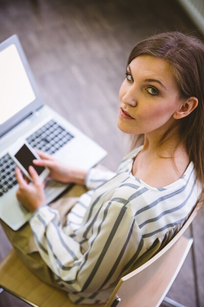 Porträt der selbstbewussten Geschäftsfrau, die Handy über Laptop im Büro verwendet