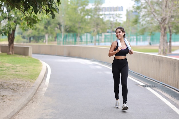 Porträt der sportlichen jungen asiatischen Frau in der Sportkleidung, die im Freien für Marathontraining rüttelt. Joggermädchen, das entlang des Betonwegs im Freien mit grünem Baumhintergrund trainiert. Sportkonzept