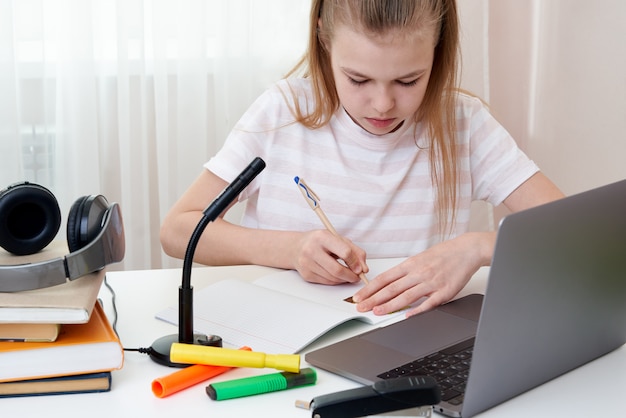 Porträt der Teenager, die online mit Kopfhörern und Laptop lernen, die Notizen in einem Notizbuch machen, das an ihrem Schreibtisch zu Hause sitzt und Hausaufgaben macht