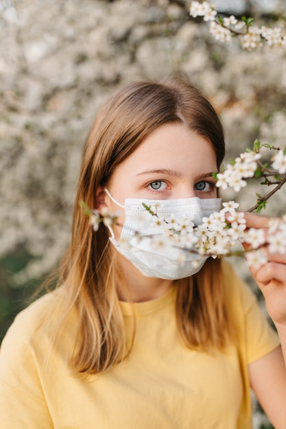 Porträt der traurigen jungen Frau in der schützenden medizinischen Gesichtsmaske mit Blumen nahe blühendem Baum in der Frühlingszeit. Frühlingsallergie-Konzept