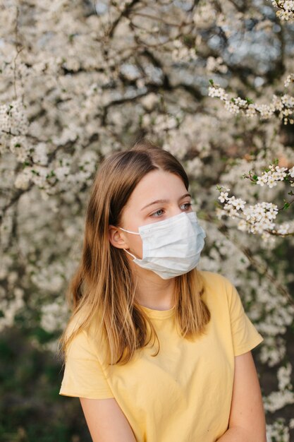 Porträt der traurigen jungen Frau in der schützenden medizinischen Gesichtsmaske mit Blumen nahe blühendem Baum in der Frühlingszeit. Frühlingsallergie-Konzept