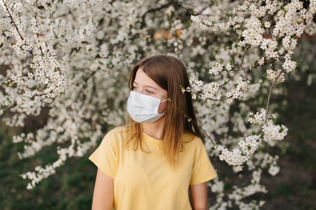 Porträt der traurigen jungen Frau in der schützenden medizinischen Gesichtsmaske mit Blumen nahe blühendem Baum in der Frühlingszeit. Frühlingsallergie-Konzept